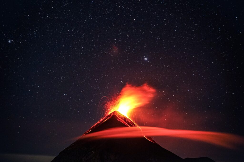 An erupting volcano is silhouetted against the starry nighttime sky. Its glow illuminates a wispy cloud near the summit.
